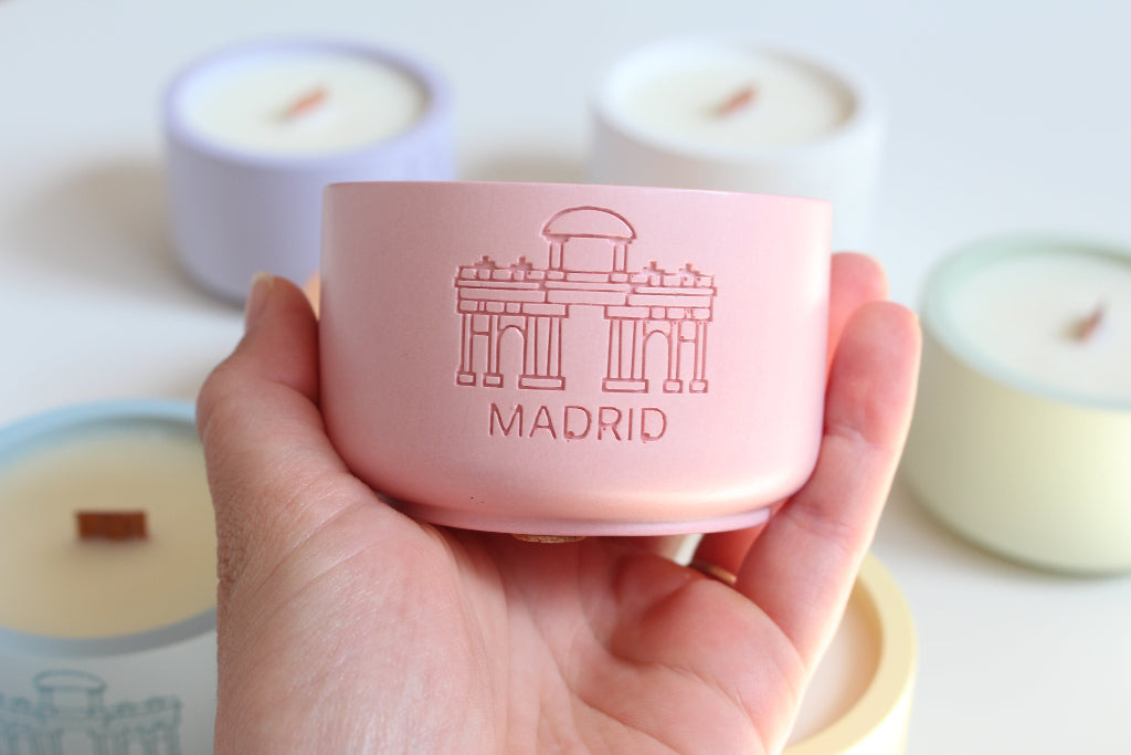Hand holding a pink container with 'Madrid' design, surrounded by candles on a light background
