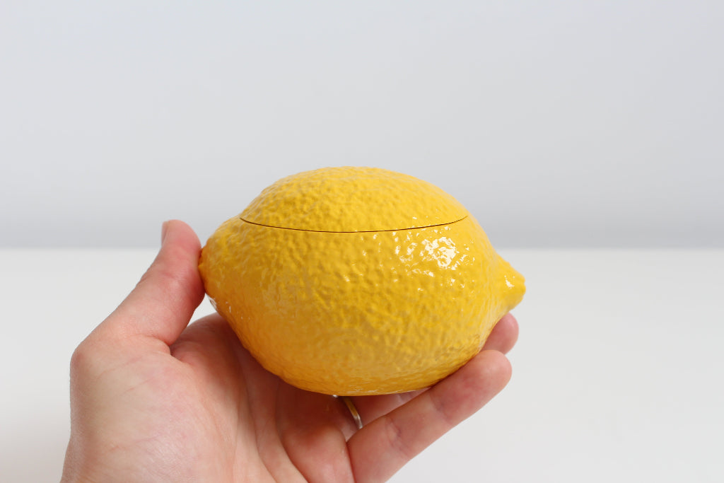 Hand holding a yellow lemon-shaped container candle against a plain background