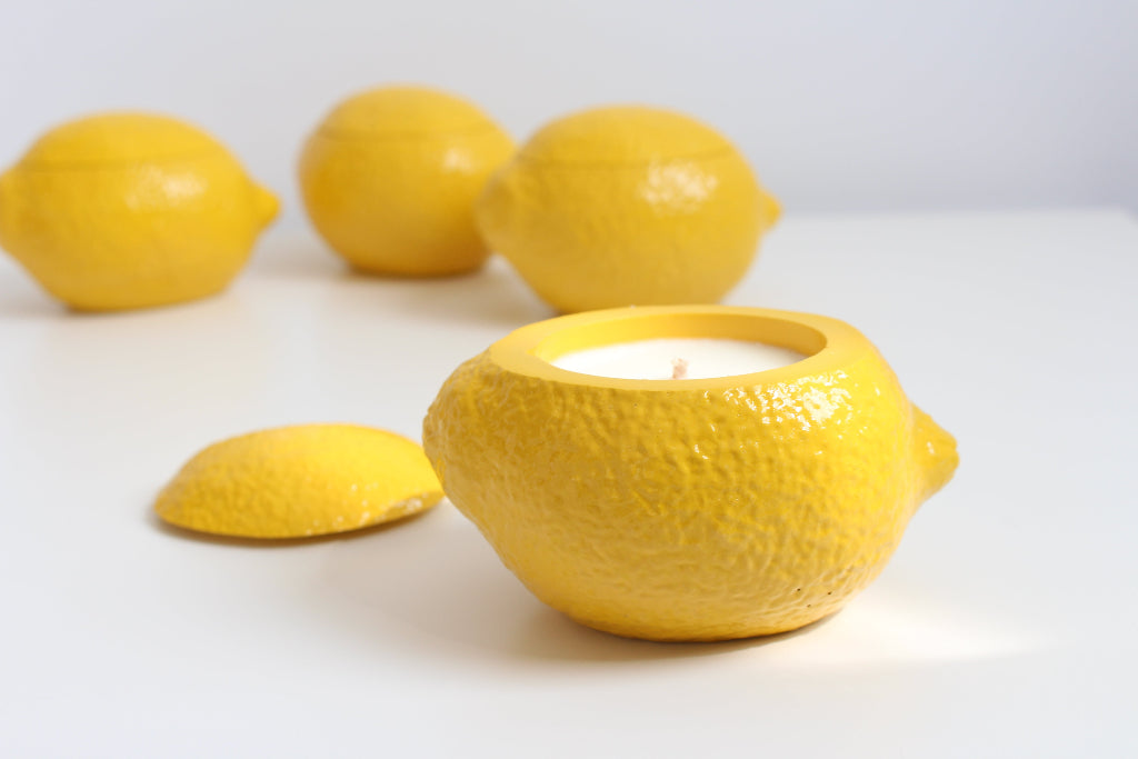 Yellow ceramic lemon-shaped candle holder with a candle inside, surrounded by real lemons on a white background.
