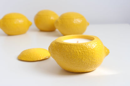 Yellow ceramic lemon-shaped candle holder with a candle inside, surrounded by real lemons on a white background.